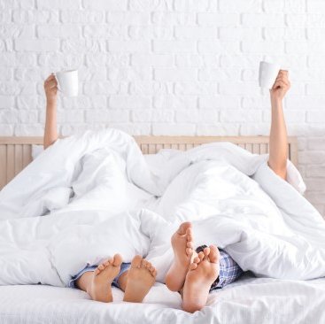 Übernachten in Tann (Rhön) - Ehepaar im Bett mit nackten Füßen halten Tasse hoch
