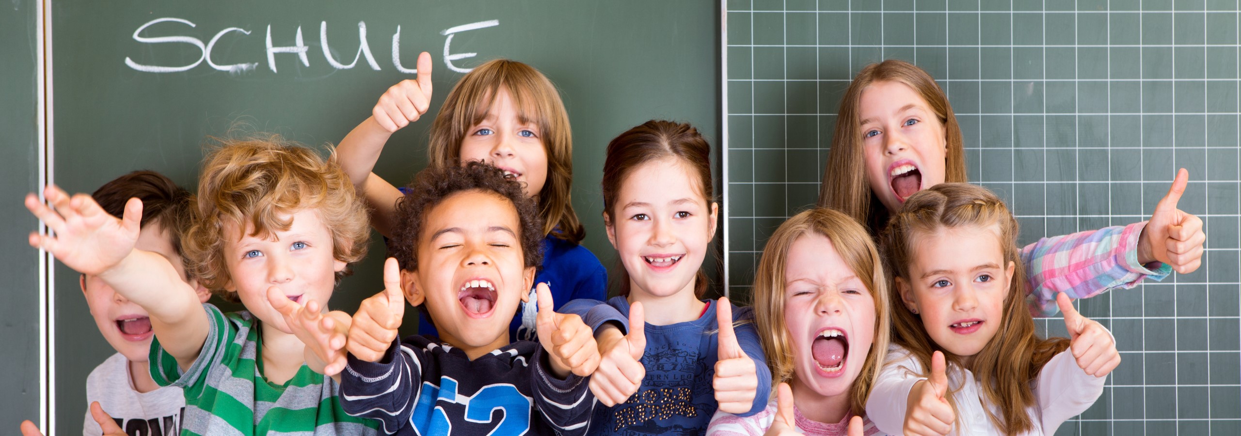 Schule - Panoramaschnitt: Jubelnde Mädchen und Jungen zeigen Daumen hoch vor einer Schultafel